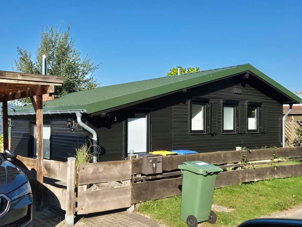 Schwarze Hütte mit grünem Dach und Holzzaun; davor steht ein grüner Mülleimer.