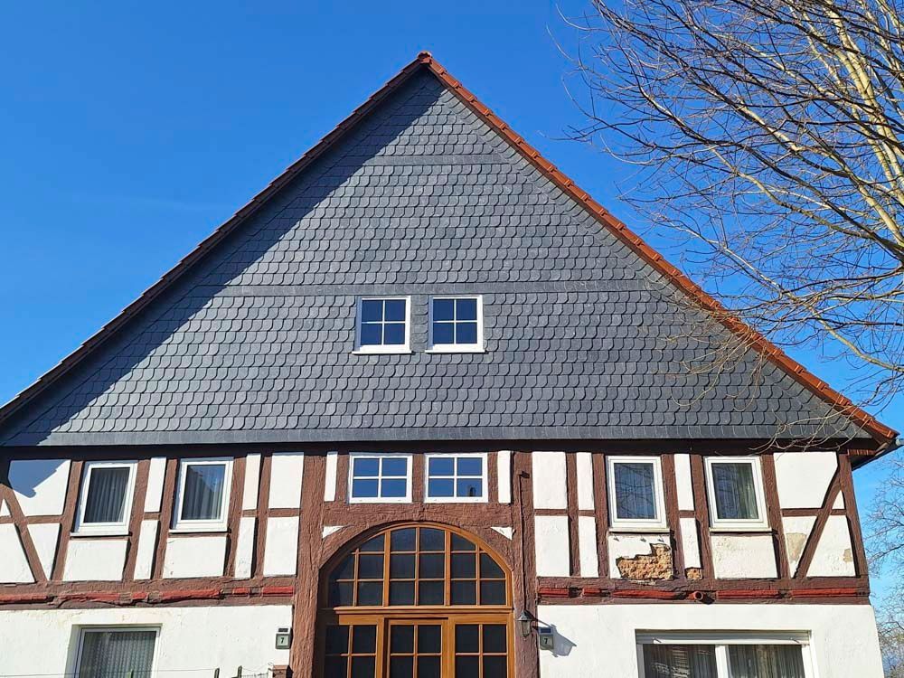 Zweistöckiges Gebäude mit grauem Schieferdach und weiß-braunem Fachwerk vor blauem Himmel.