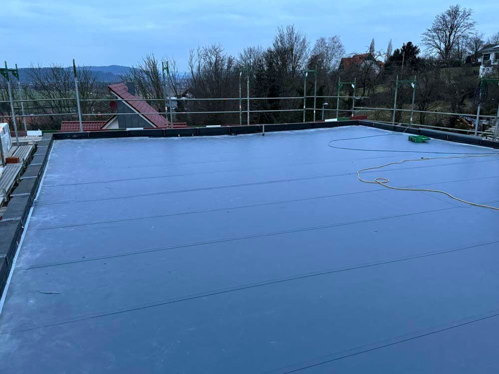 Flaches, dunkelgraues Dach mit Gerüst am Rand, im Hintergrund Bäume und Gebäude.