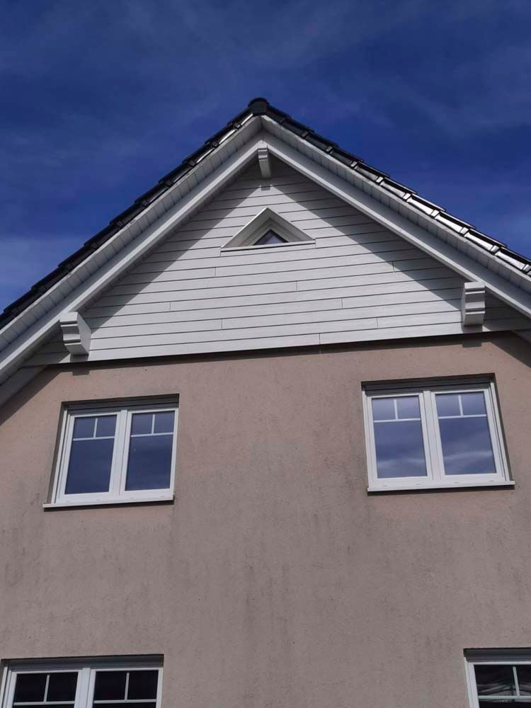 Beige Hausfassade mit weißen Zierelementen, zwei Fenstern und einem dreieckigen Giebel unter blauem Himmel.