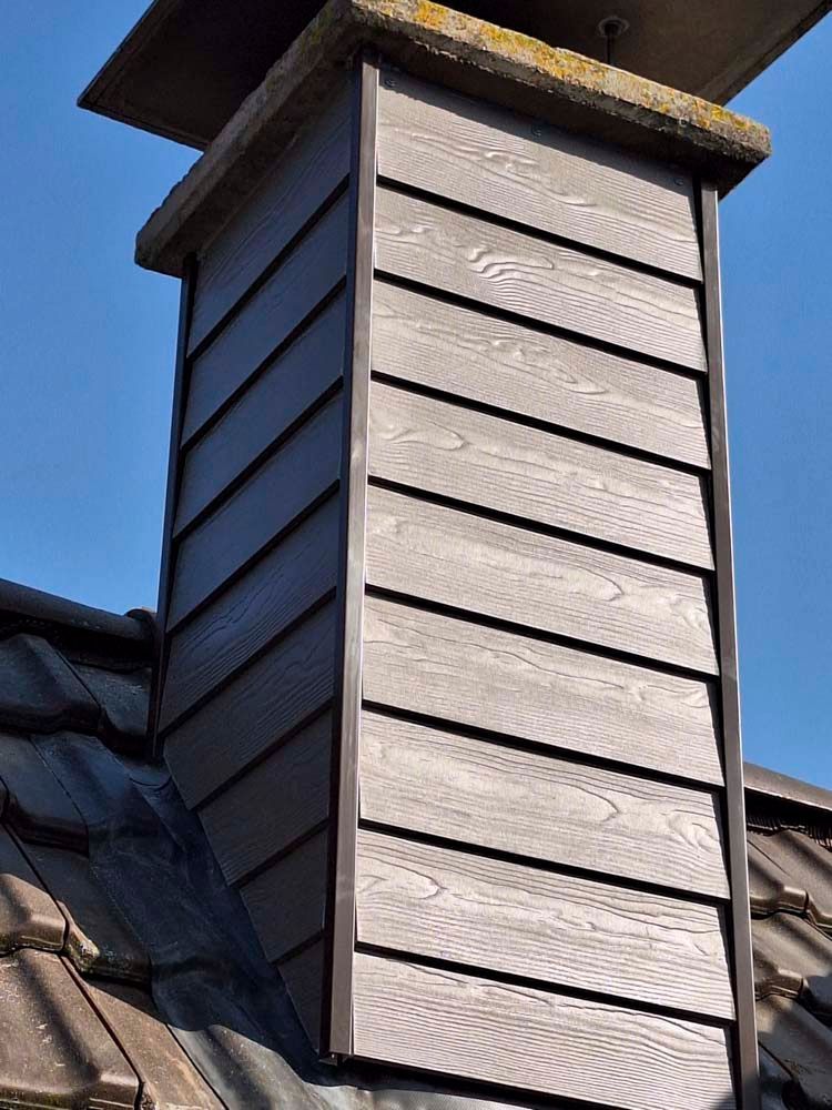Ein Schornstein auf einem Dach, verkleidet mit braunem, strukturiertem Holz, mit einer Betonplatte und dunklen Zierelementen.