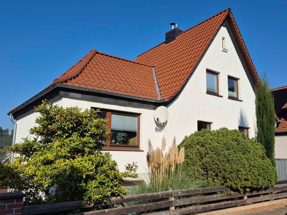 Weißes zweistöckiges Haus mit rotem Ziegeldach, umgeben von grünen Sträuchern und einem Holzzaun, unter blauem Himmel.