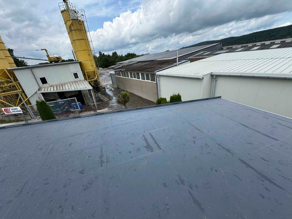 Industriekomplex: graues Dach im Vordergrund, gelbes Silo, Gebäude, bewölkter Himmel.