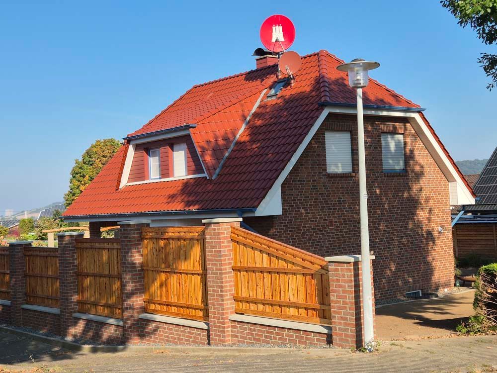 Ein Backsteinhaus mit rotem Dach, Holzzaun, roter Satellitenschüssel und Straßenlaterne an einem sonnigen Tag.