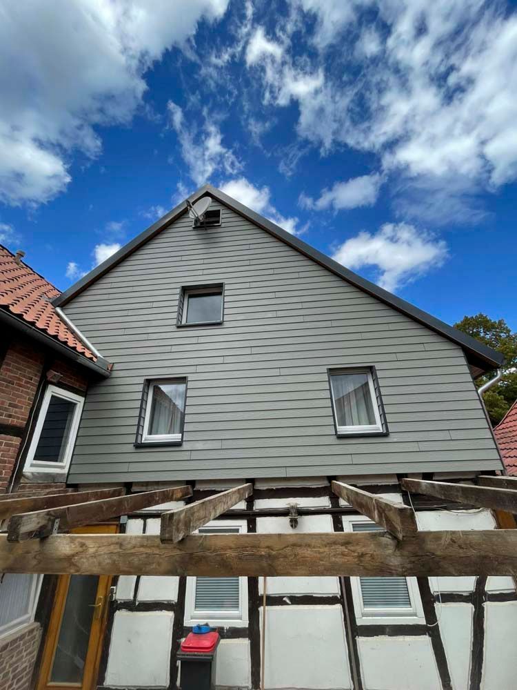 Ein zweistöckiges Haus mit dunklem Schieferdach und weißen Stuckwänden. An das Haus ist eine hölzerne Pergola angebaut.