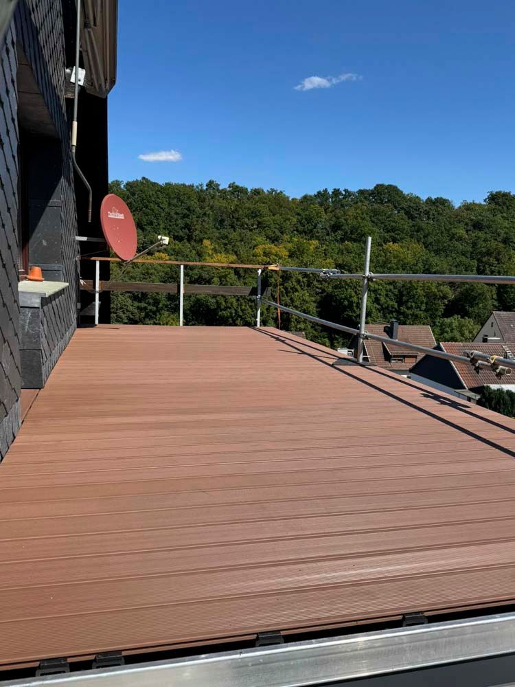 Holzterrasse auf dem Dach mit Metallgeländer, Satellitenschüssel und Blick auf grüne Bäume unter blauem Himmel.