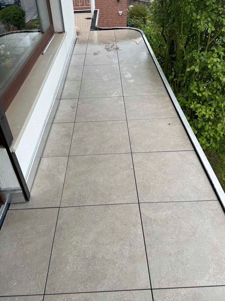 Grau gefliester Balkon mit Fenster links und Bäumen rechts.