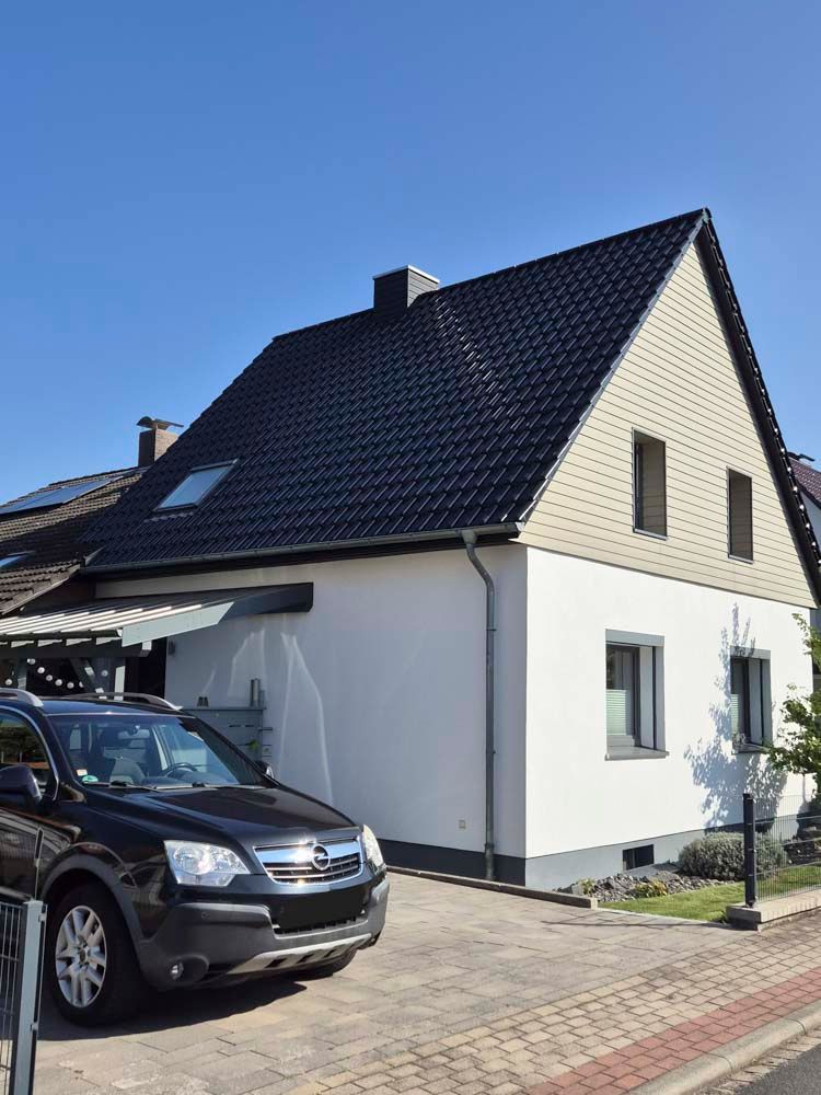 Weißes Haus mit schwarzem Dach, geparktes Auto und blauer Himmel.