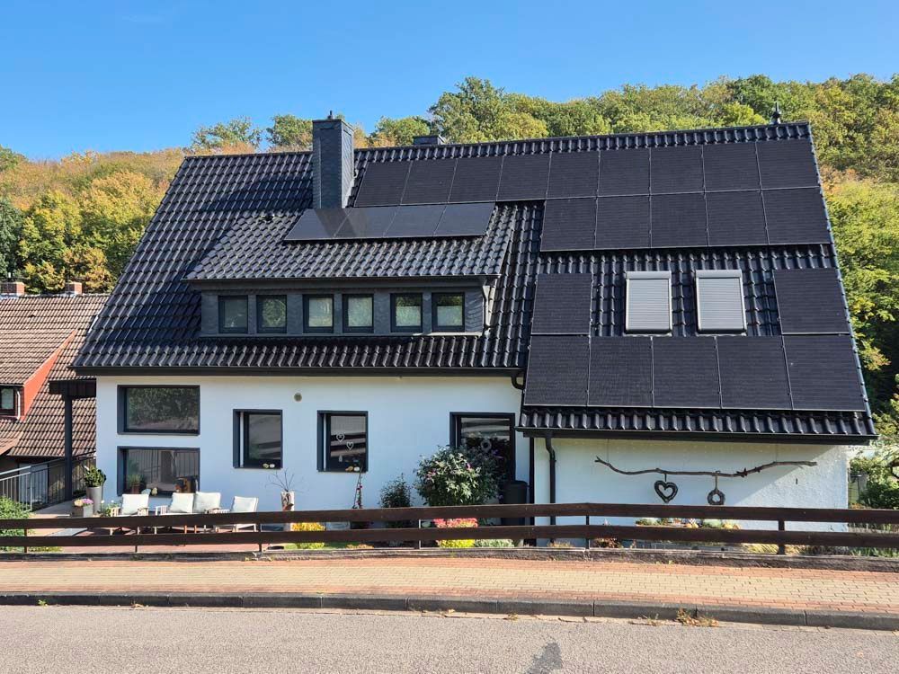 Weißes Haus mit schwarzem Ziegeldach, das mit Solarpaneelen bedeckt ist, mit Dachgauben und grünem Laub im Hintergrund.