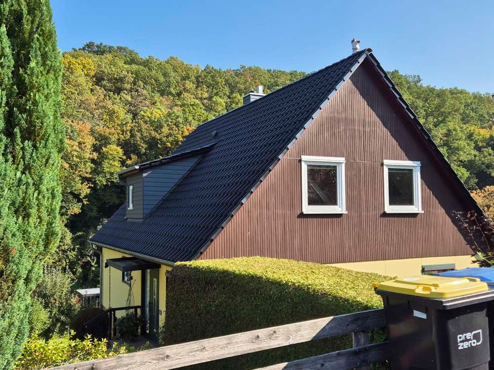 A-förmiges Haus mit brauner Fassade, schwarzem Dach und zwei Fenstern. Im Hintergrund grüne Hecke und Bäume.