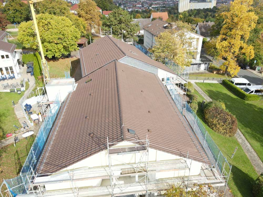 Gebäude mit braunem Ziegeldach, Gerüst und Sicherheitsnetzen, umgeben von grünen Rasenflächen und Bäumen.