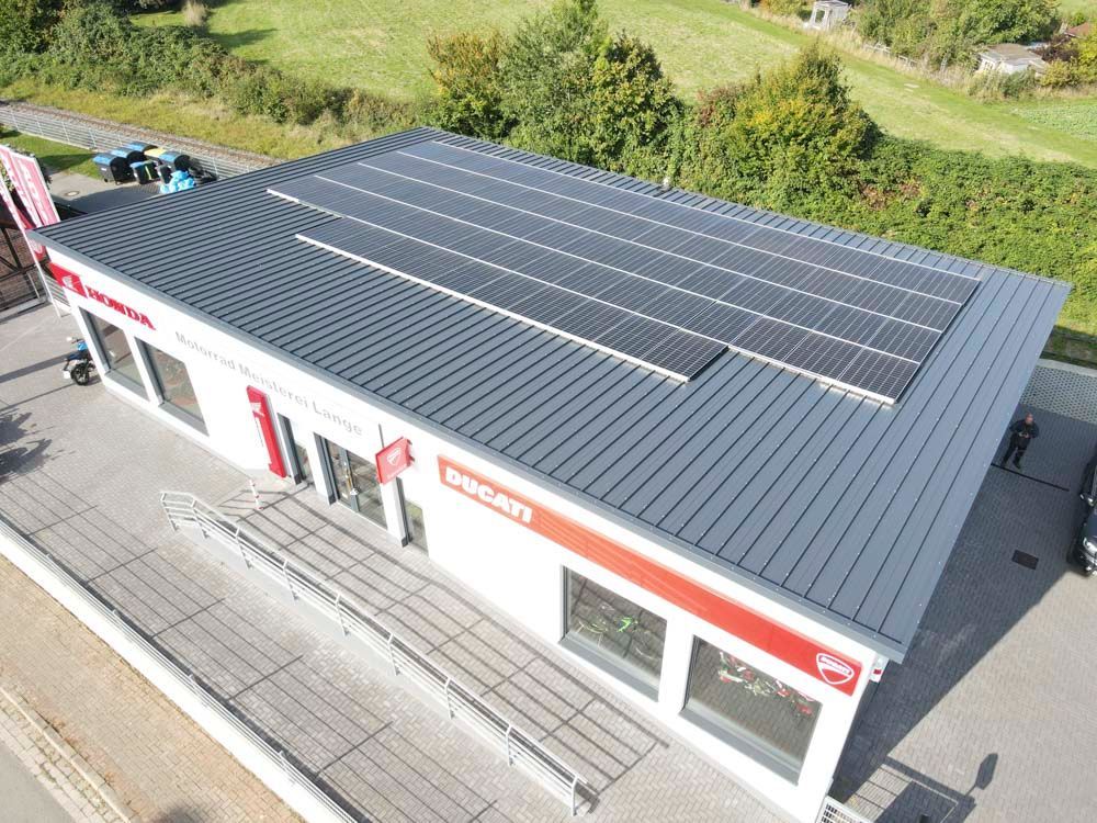 Geschäft mit grauem Dach und Solarpaneelen, rotem Logo und Rampe; Auto in der Nähe geparkt.