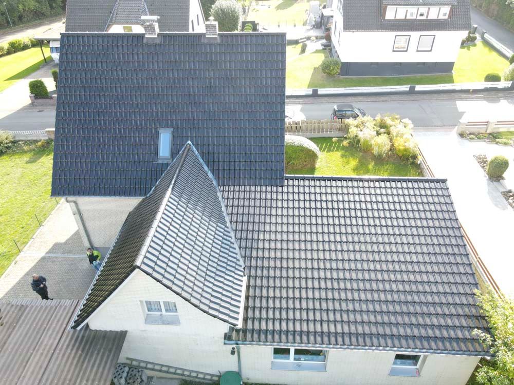 Draufsicht auf ein Haus mit dunkelgrauem Ziegeldach, einen Rasen und eine Straße in sonniger Lage.