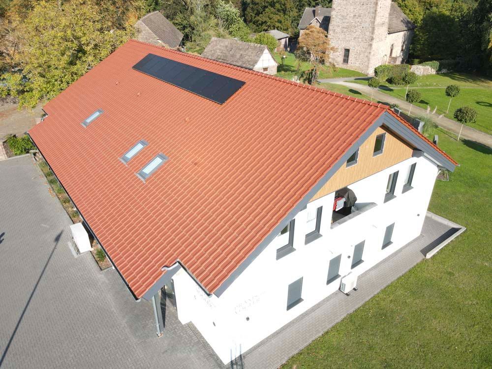Weißes Gebäude mit rotem Ziegeldach, Solarpaneelen und kleinen Fenstern auf einem grünen Rasen.