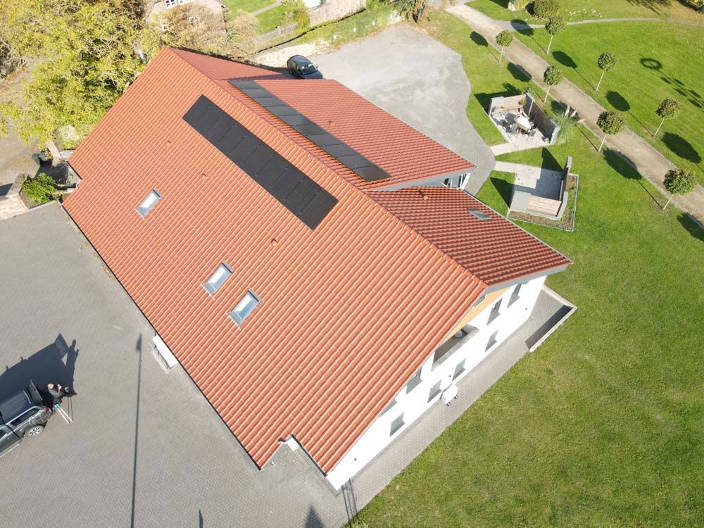 Luftaufnahme eines weißen Gebäudes mit orangefarbenem Ziegeldach, Solarpaneelen und Dachgauben, auf einem grünen Rasen.