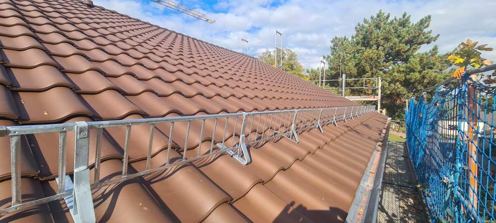 Braunes Ziegeldach mit metallenem Sicherheitsgeländer, blauer Himmel und Bäume im Hintergrund.