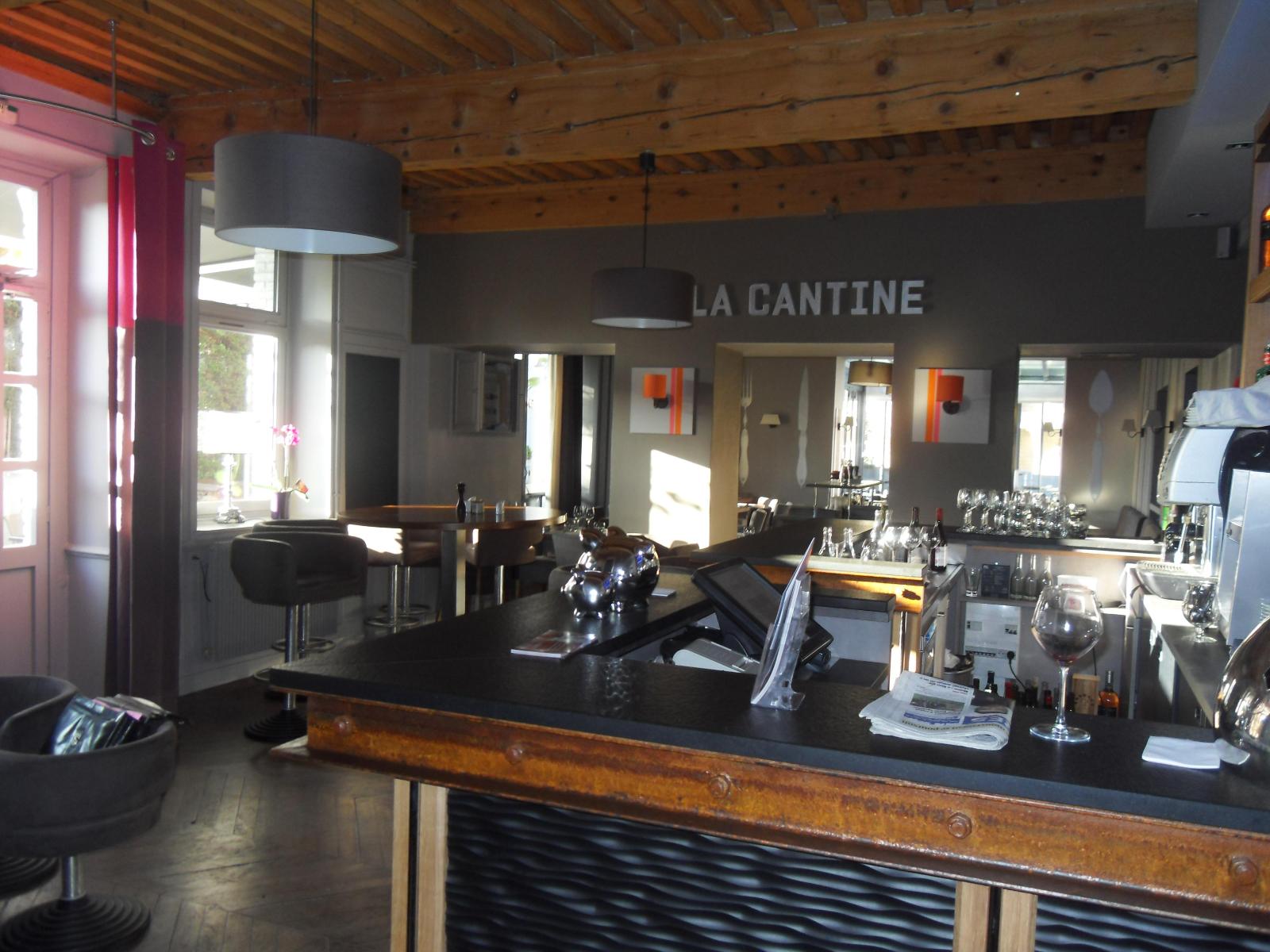 Vue intérieure d'un bistro nommé La Cantine, avec son comptoir en bois, ses chaises sombres et ses suspensions lumineuses minimalistes.