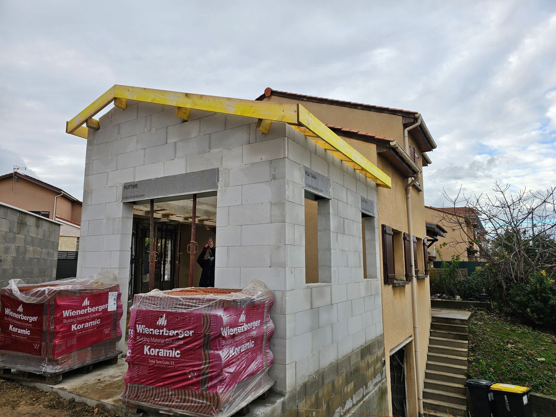 Une extension inachevée en parpaings, attenante à une maison beige de deux étages, avec deux palettes de briques emballées en rouge à proximité.