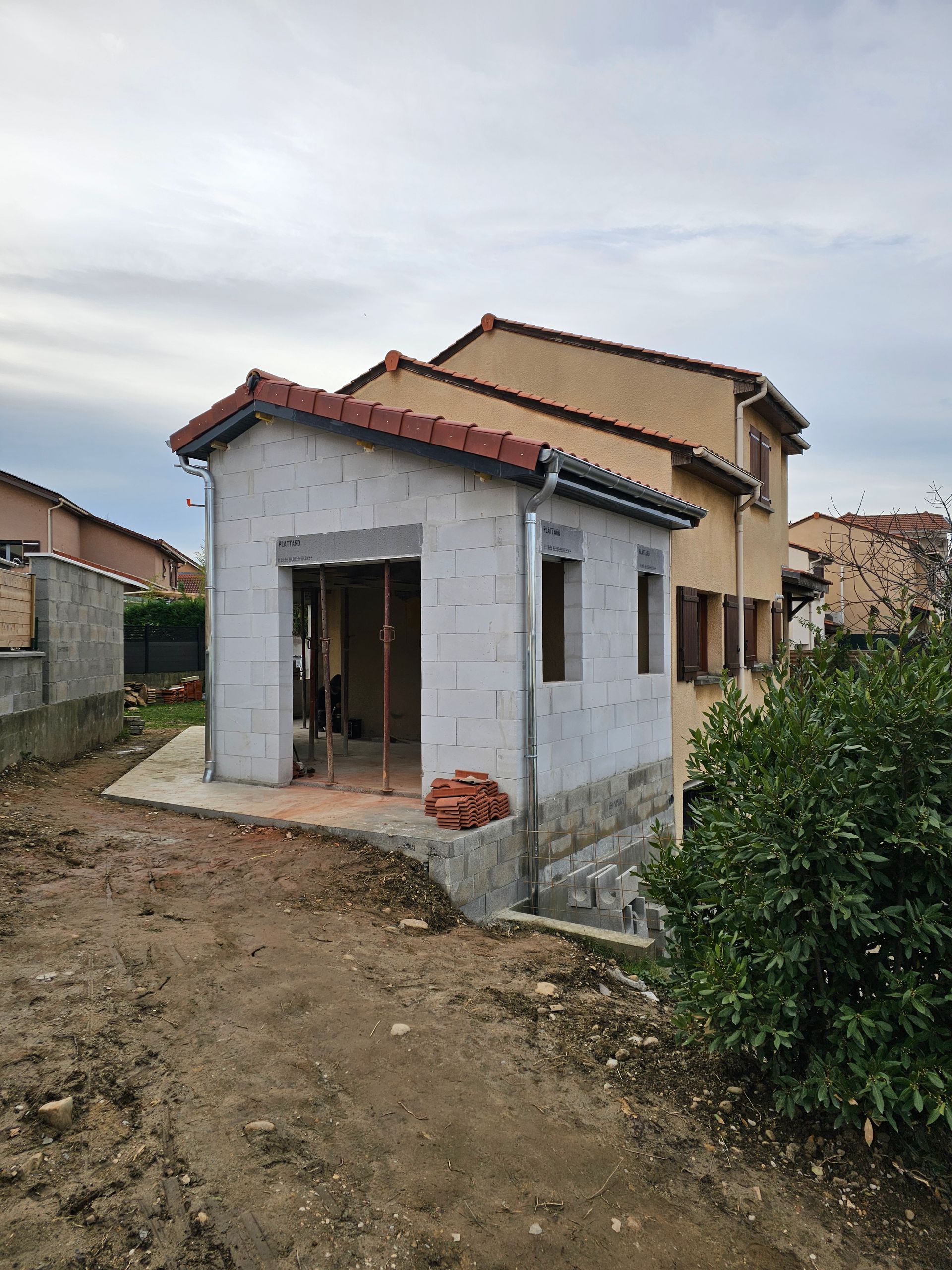 Une extension en béton clair, récemment construite, est adossée à une maison beige de deux étages, sous un ciel nuageux.