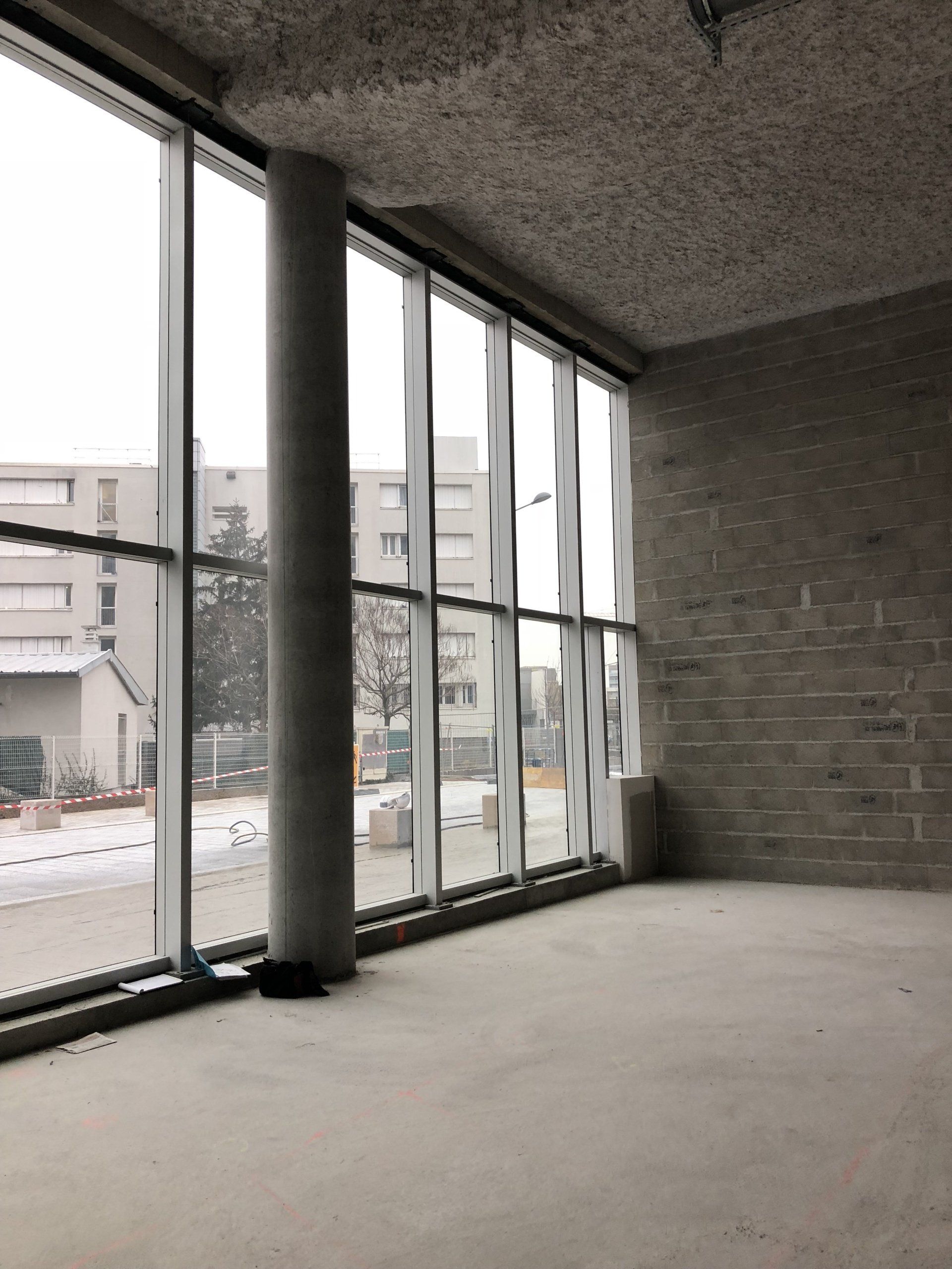 Une pièce vide aux sols et murs en béton brut, dotée de baies vitrées donnant sur une rue et des bâtiments.