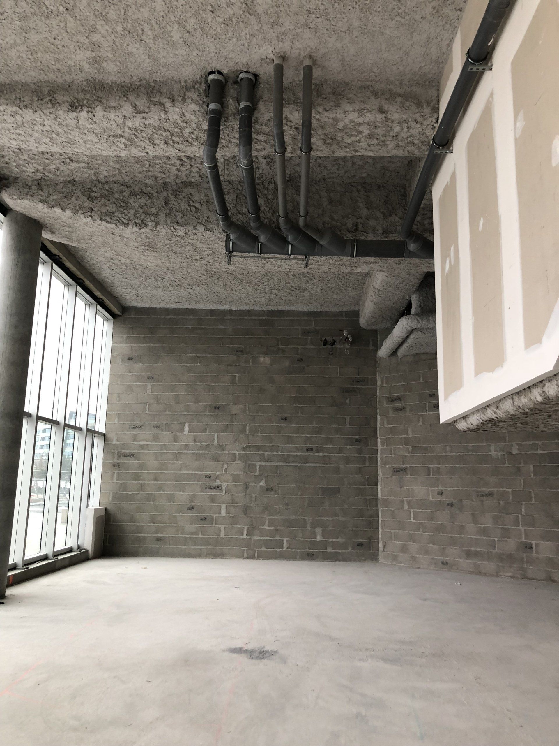 Un espace intérieur inachevé avec des murs gris texturés, des tuyaux apparents au plafond en béton et une grande baie vitrée.