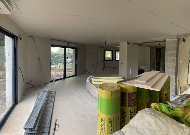 Intérieur d'une maison en construction avec des murs blancs, des portes coulissantes en verre et des rouleaux de matériau isolant jaune.