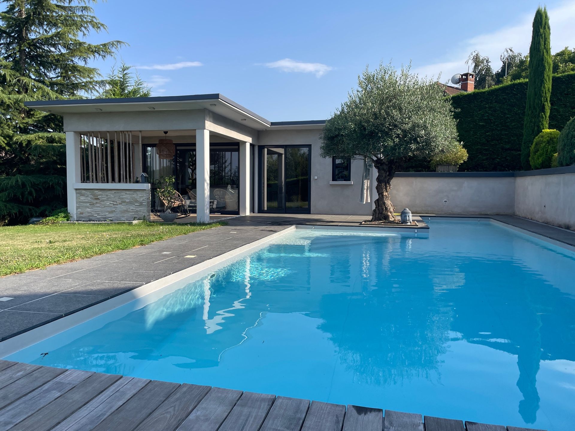 Par une journée ensoleillée, une piscine aux eaux bleues se trouve devant une maison moderne dotée d'un patio couvert et d'un petit arbre.