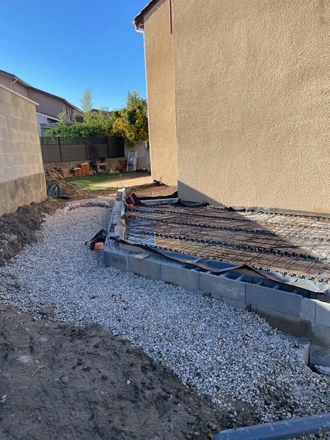 Chantier de construction montrant les fondations d'une maison avec des bordures en parpaings, un treillis métallique et un aménagement paysager en gravier.
