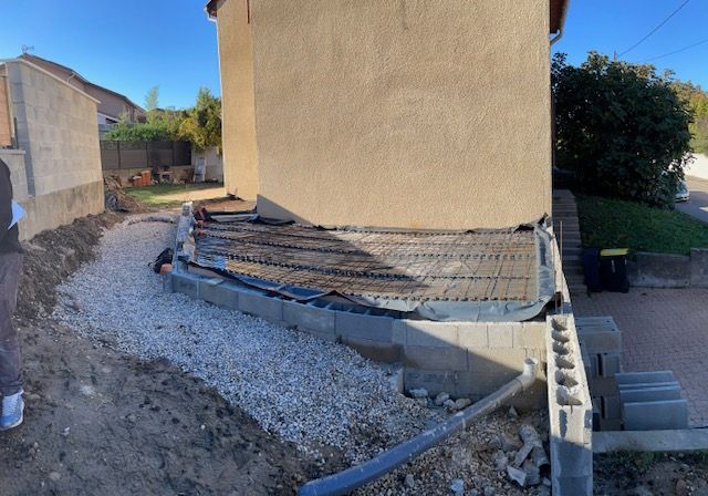 Une structure de patio en béton partiellement construite contre une maison, avec du gravier, des parpaings et un treillis d'armature métallique.