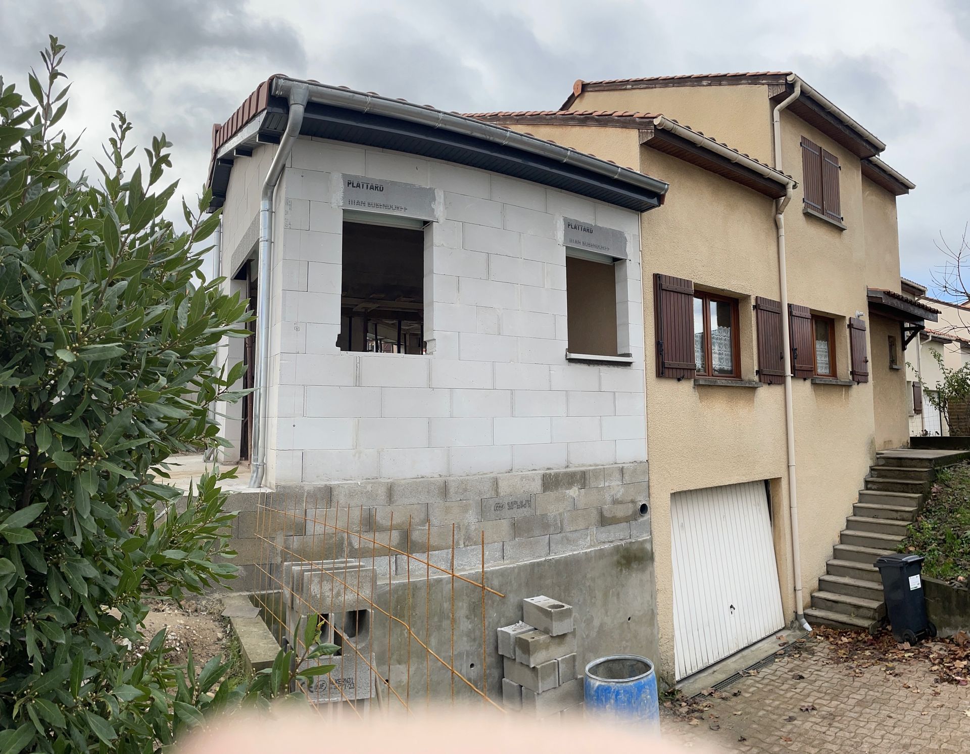 Une maison beige à deux étages avec une extension inachevée aux murs de parpaings blancs sur le côté gauche et un garage en dessous.