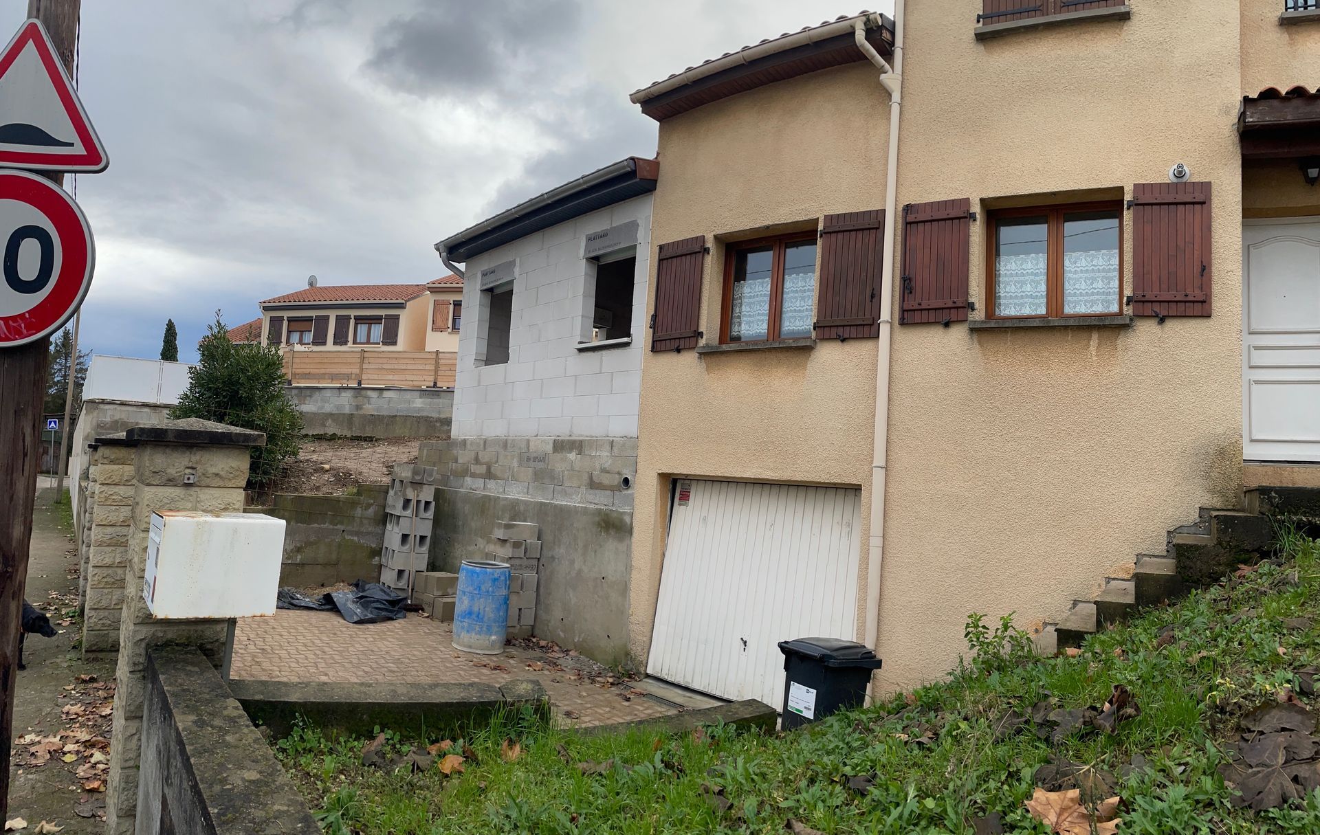 Vue de rue d'une maison beige à deux étages avec des volets marrons et un garage blanc, à côté d'un bâtiment blanc plus petit.