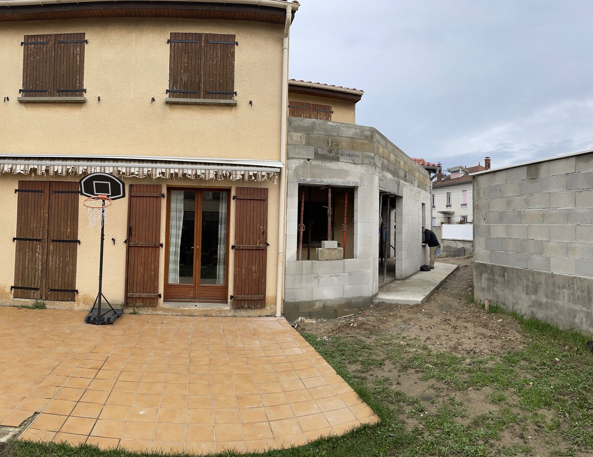 Une maison beige à deux étages avec des volets marron se trouve à côté d'une extension en parpaings en construction dans une cour.
