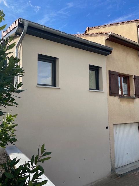 Façade d'un bâtiment couleur crème avec deux fenêtres et une gouttière, situé à côté d'une maison plus ancienne avec un garage.