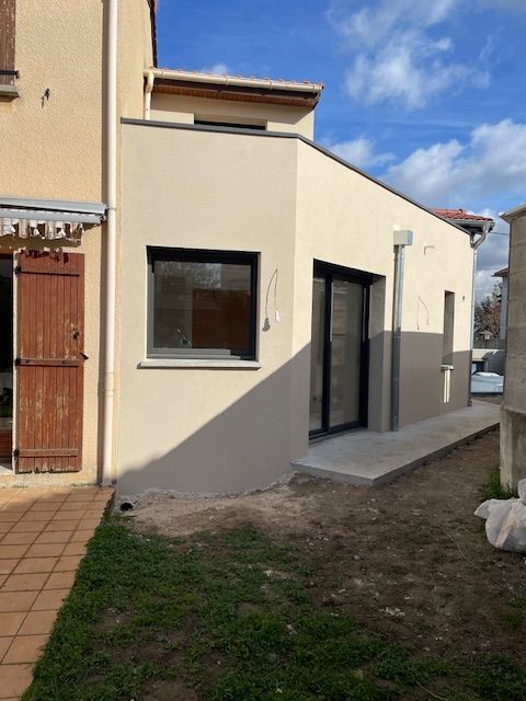 Une extension moderne, de couleur beige clair, avec de grandes fenêtres et une porte coulissante en verre, jouxte un bâtiment plus ancien.