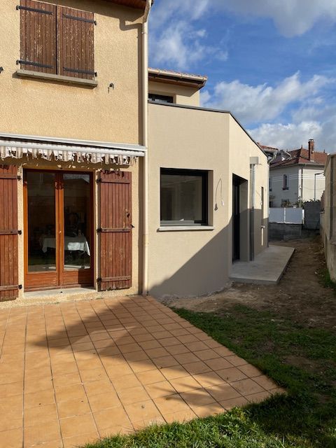 Une terrasse carrelée et une pelouse devant une maison beige de deux étages avec une extension plus récente à toit plat et des volets en bois.