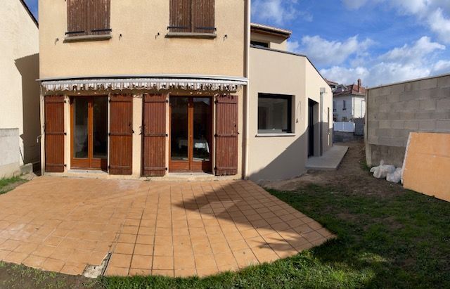 Une maison beige avec deux portes-fenêtres, des volets en bois et une terrasse carrelée, donnant sur une cour latérale non pavée.