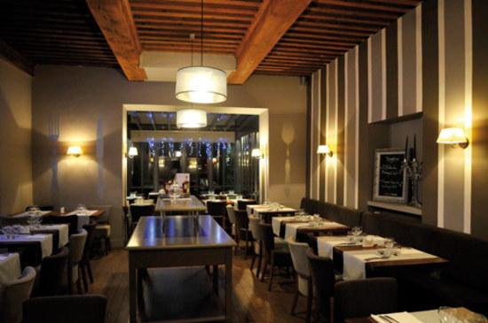 Un intérieur de restaurant chaleureux et tamisé, avec des poutres apparentes au plafond, des murs rayés, des appliques murales aux tons chauds et des tables à manger disposées avec soin.