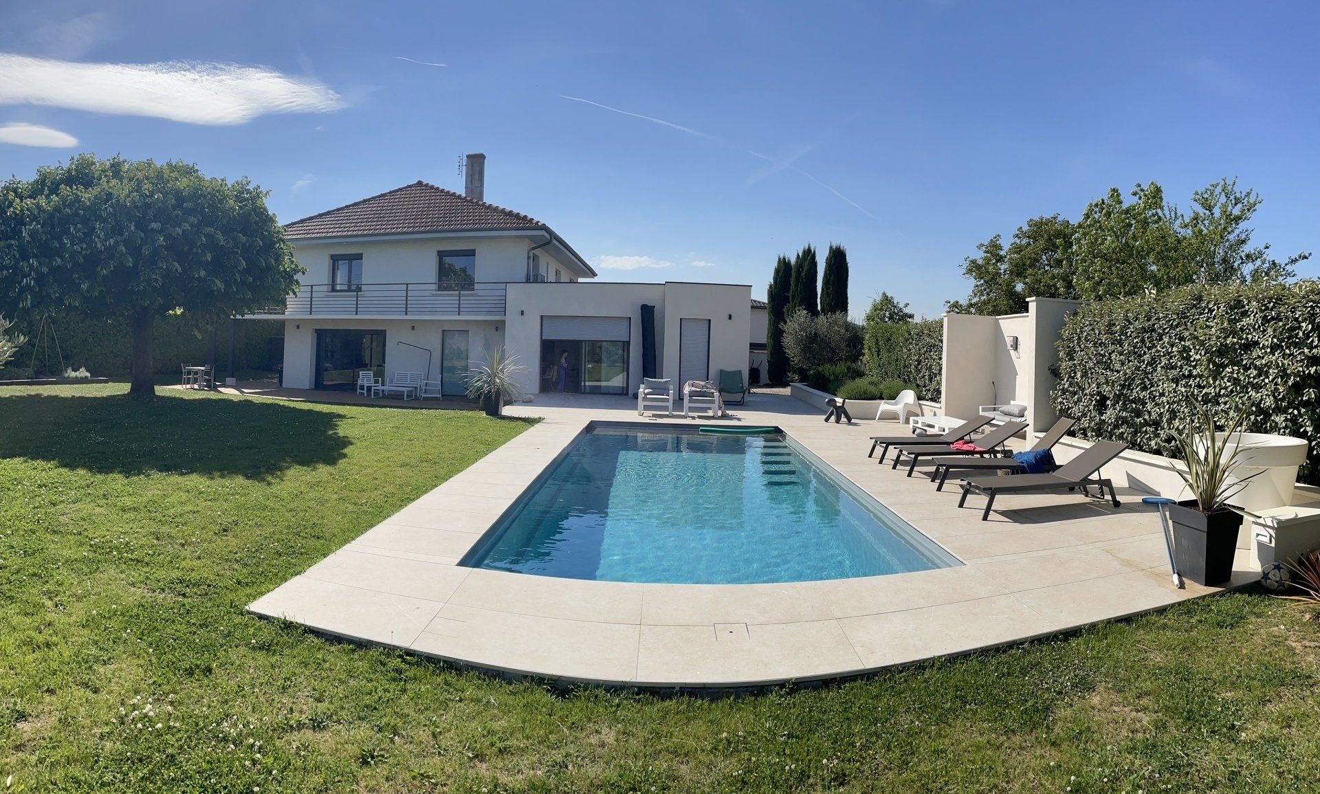 Une maison à deux étages avec une piscine rectangulaire bleue sur une terrasse, entourée d'une pelouse sous un ciel bleu azur.