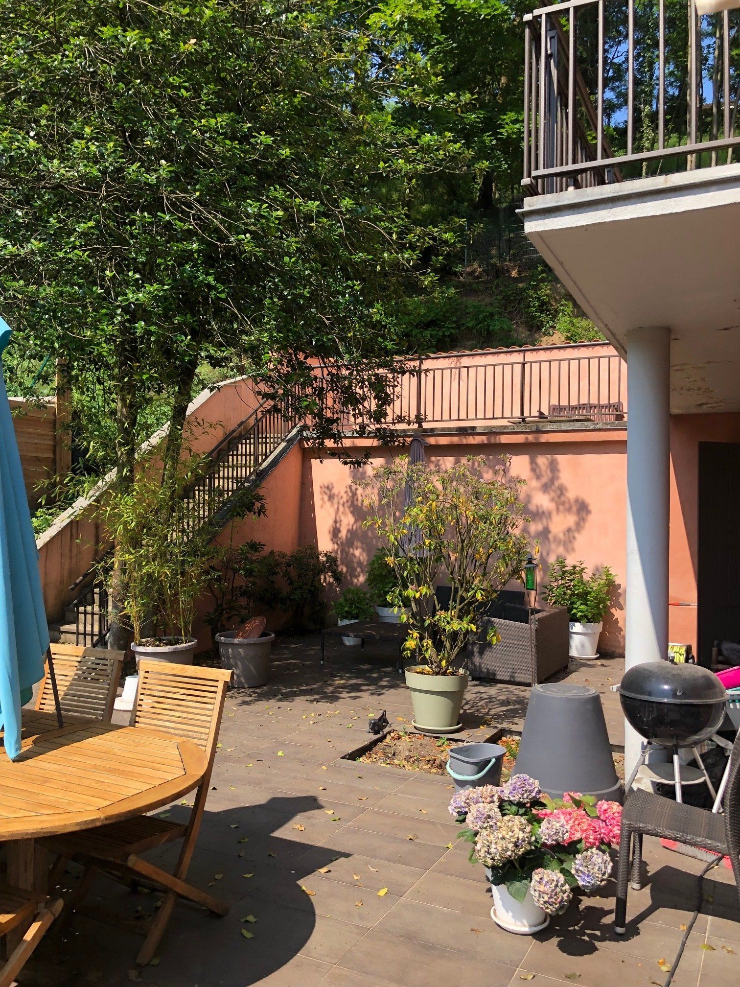 Un patio ensoleillé avec une table ronde en bois, des plantes en pot, un petit barbecue au charbon de bois et un escalier en pierre menant à une terrasse.
