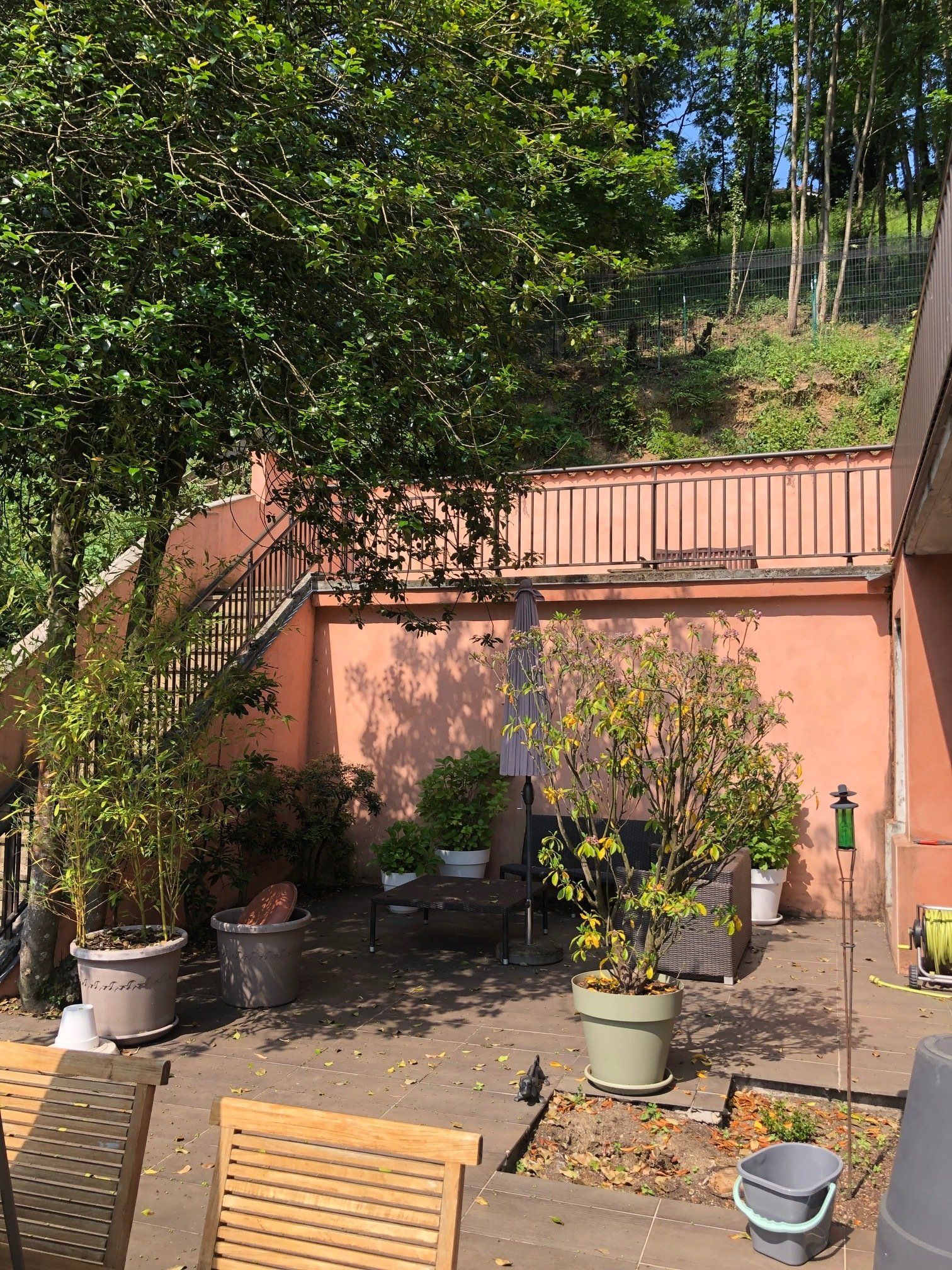 Une terrasse ensoleillée avec des plantes en pot, une petite table de bistrot et un muret à plusieurs niveaux couleur saumon menant à flanc de colline.