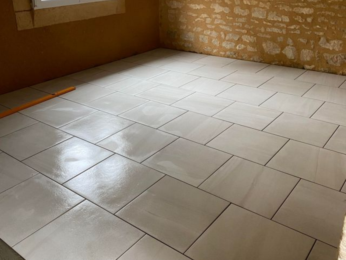 Sol carrelé blanc en cours de pose dans une pièce aux murs beiges et un mur en pierre.