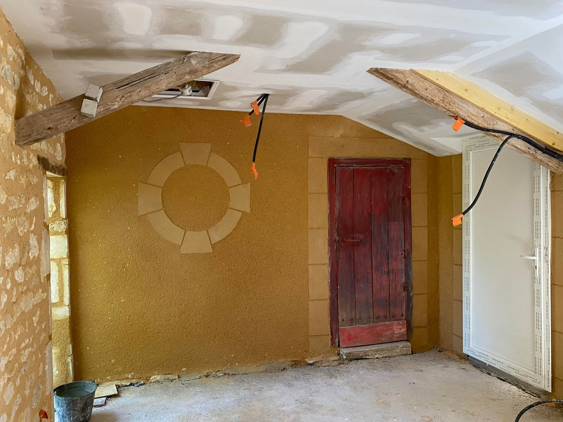 Rénovation intérieure de la pièce en cours : mur jaune texturé avec poutres en bois et une porte rouge.