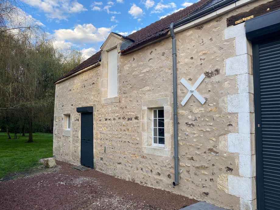Bâtiment en pierre avec une porte sombre, une fenêtre blanche et un marquage en forme de X ; arbres et ciel bleu en arrière-plan.