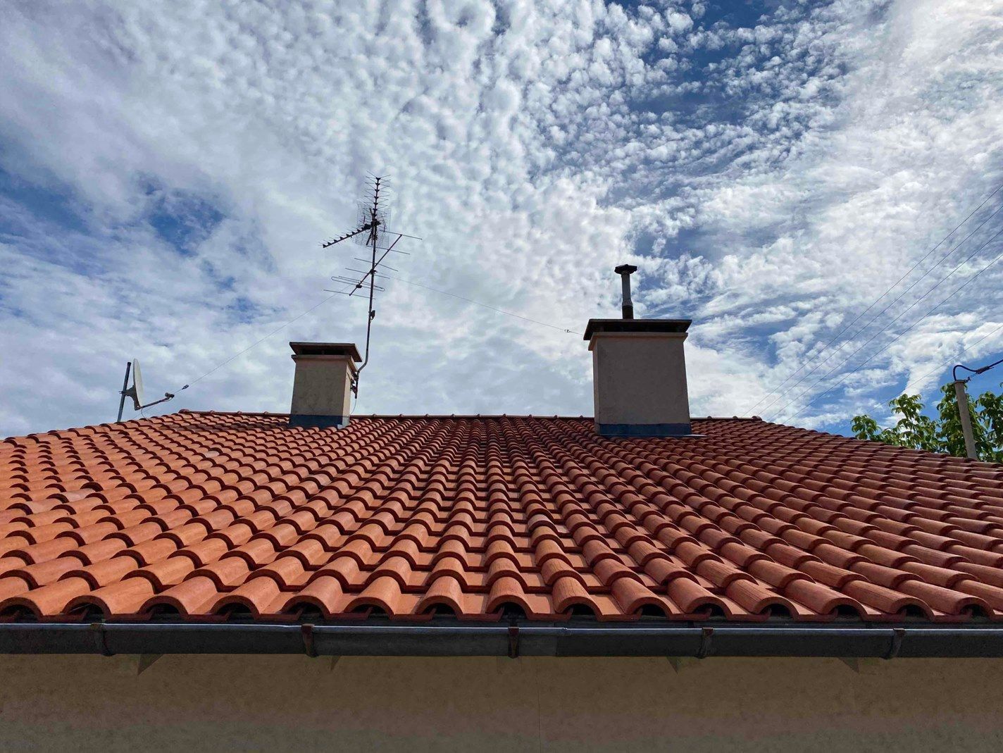 Toit de tuiles rouges avec des cheminées sous un ciel bleu partiellement nuageux, après un démoussage