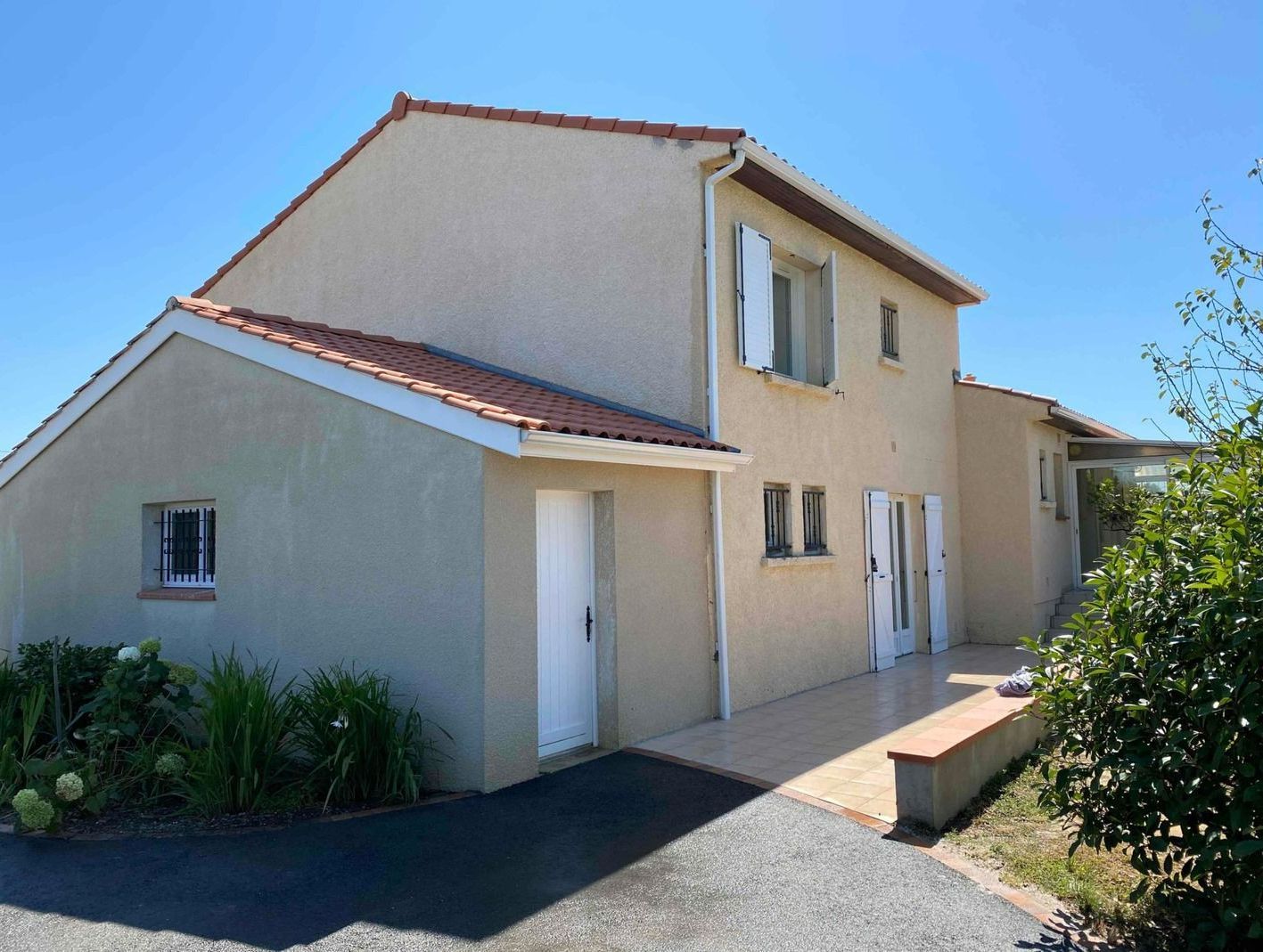 Une maison beige à deux étages avec un toit en pente, des portes blanches et une allée sous un ciel bleu clair
