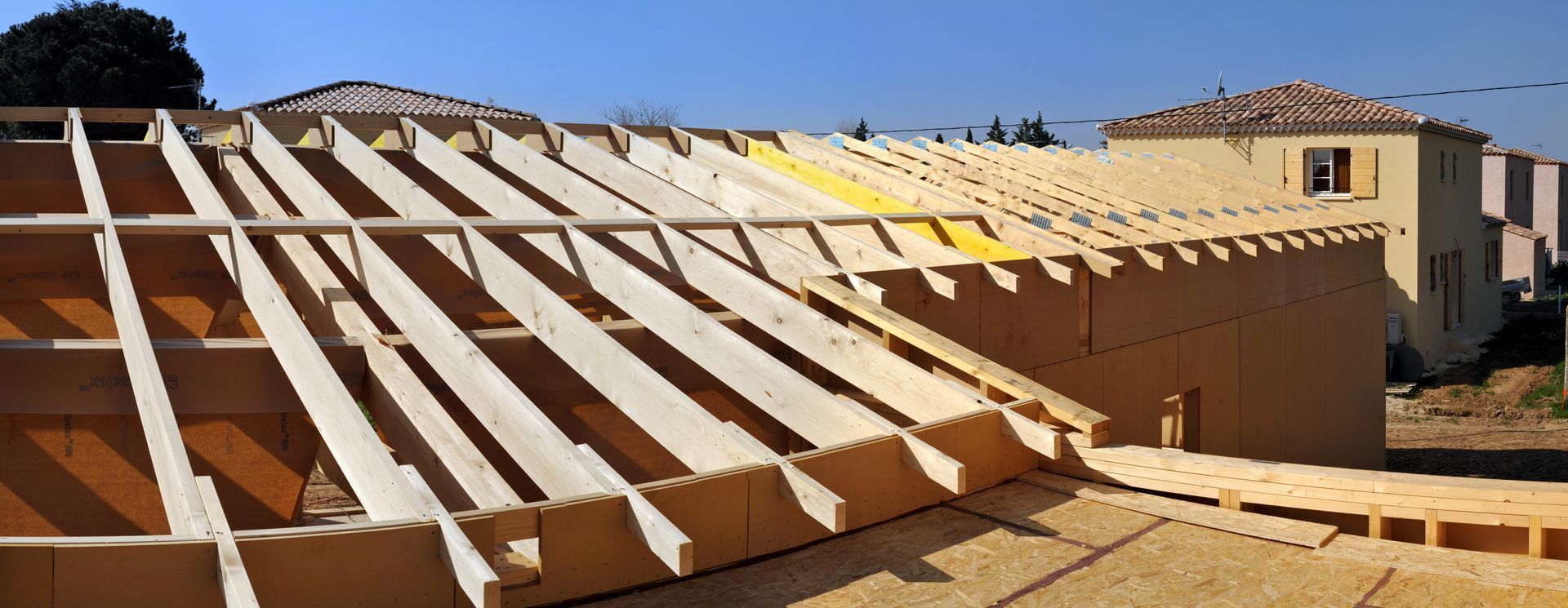 Charpente de toit en bois partiellement construite sur un bâtiment beige sous un ciel bleu clair