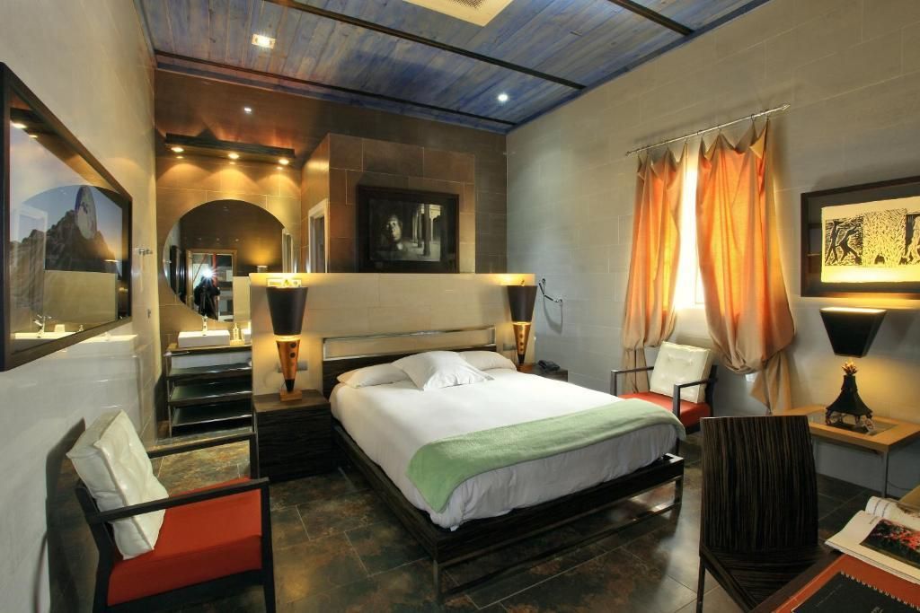 Habitación de hotel con cama, sillas y obras de arte. Cortinas naranjas y doradas, techo azul.