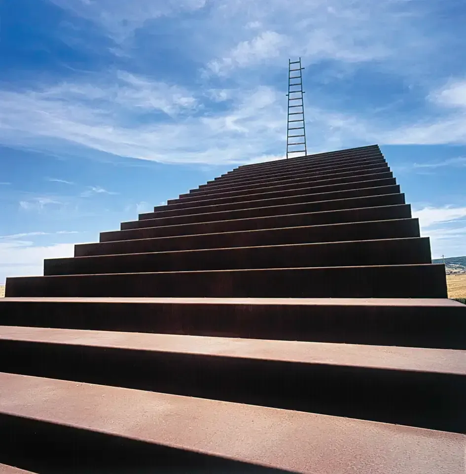 Gran escalera de metal oxidado que conduce a una escalera alta contra un cielo azul con nubes dispersas.