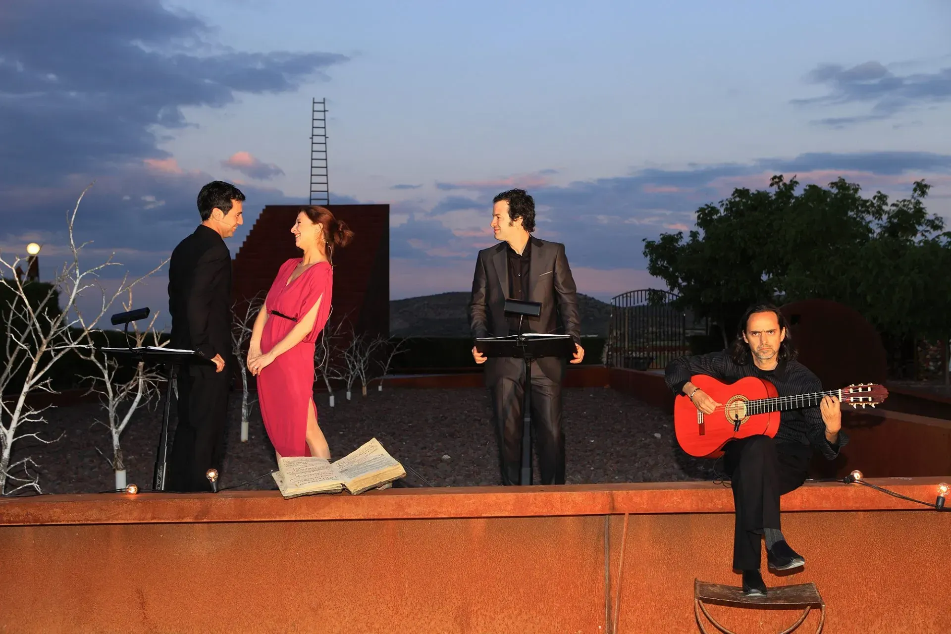 Músicos tocando en una azotea al anochecer. Un guitarrista está sentado, mientras otros, de pie, leen partituras, vestidos de gala.