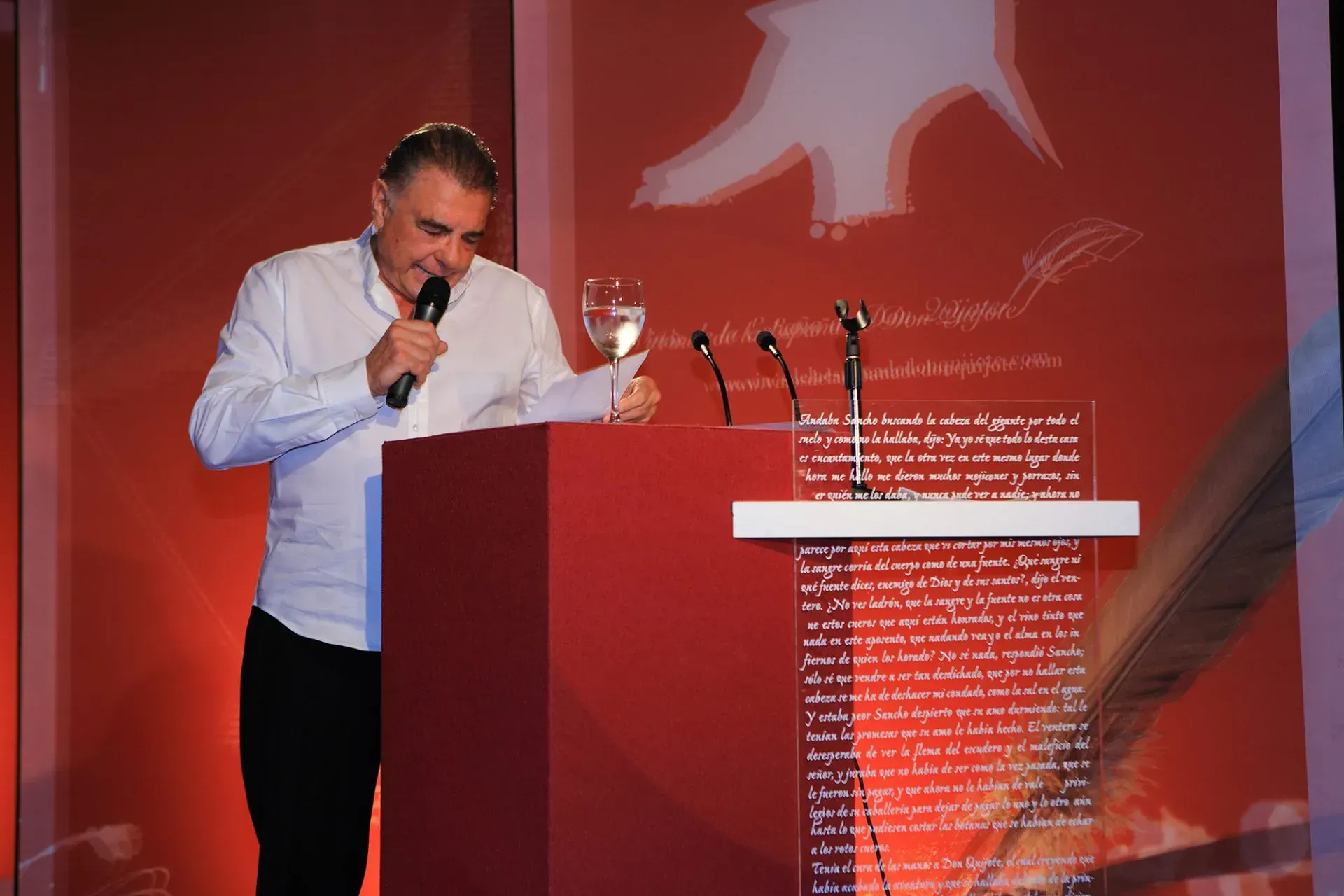 Hombre en un podio con un micrófono y un vaso de agua, hablando en un evento con fondo rojo.